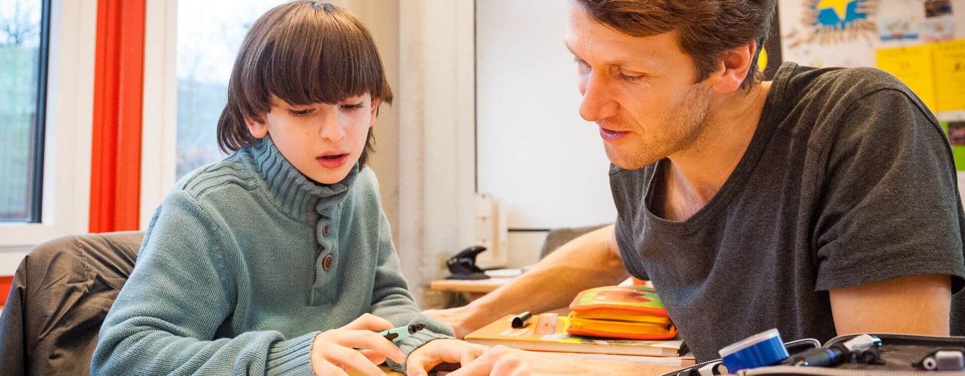 Lehrkraft und Schüler sitzen an einem Tisch in der Stadtteilschule Mümmelmannsberg, umgeben von Schulmaterialien und bunten Wandpostern.